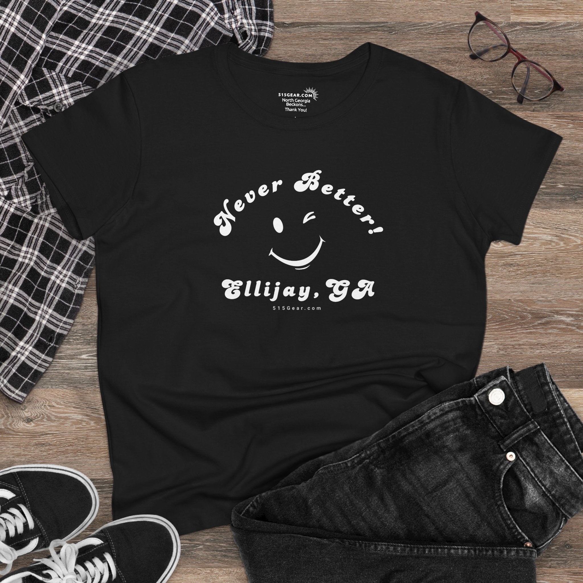 Black t-shirt with 'Never Better! Ellijay, GA' text and smiley face on a wooden surface with jeans and sneakers.