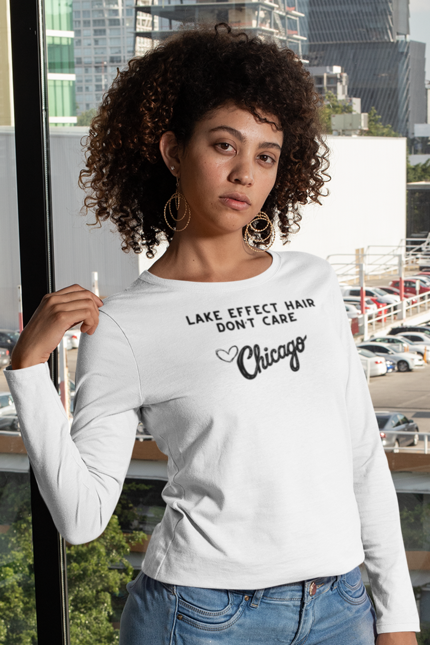 Woman wearing a white long sleeve shirt with text "Lake Effect Hair Don't Care... Love Chicago" standing indoors with a cityscape in the background