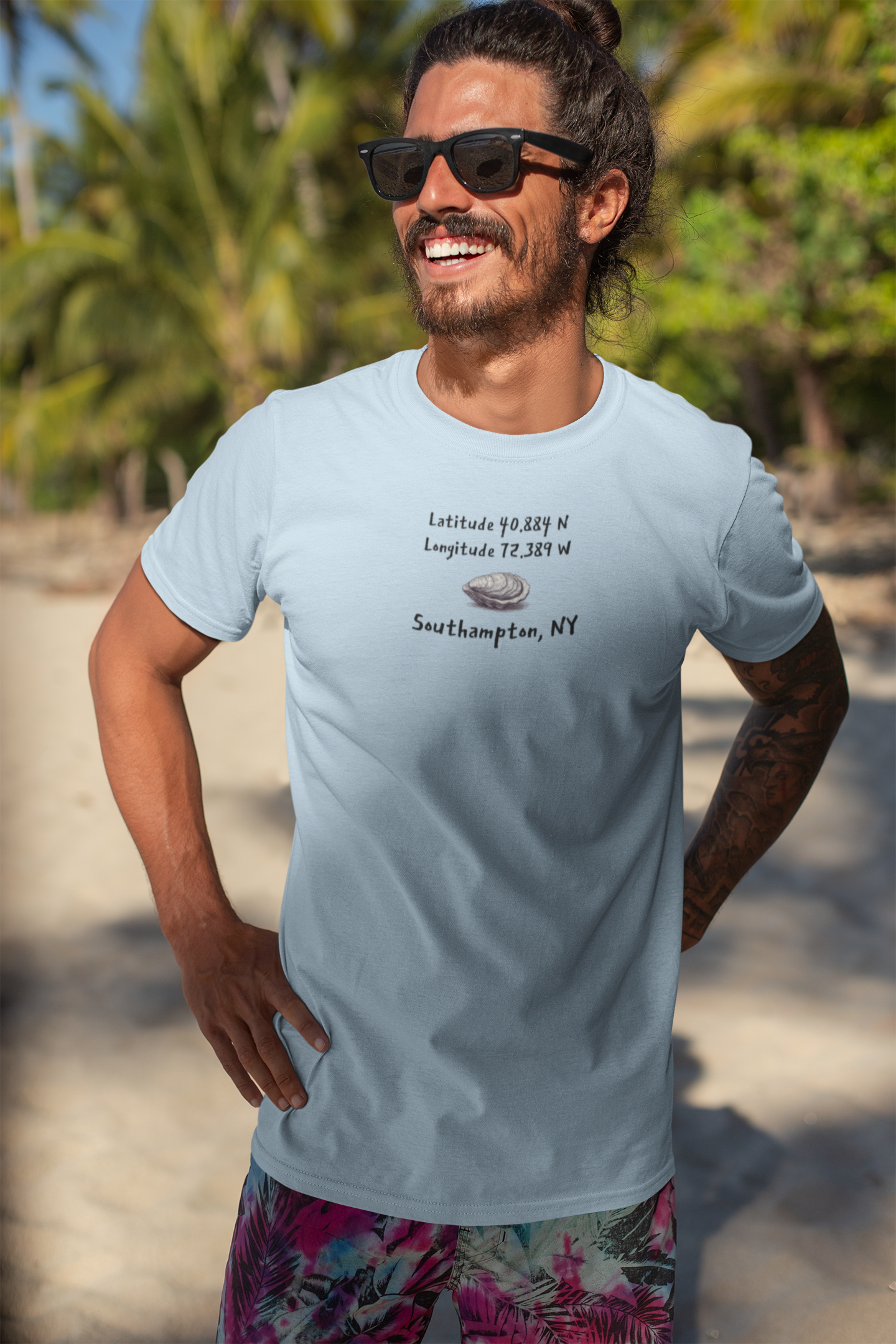 Man wearing a light blue t-shirt with oyster graphic and latitude and longitude for Southampton NY standing on a beach.