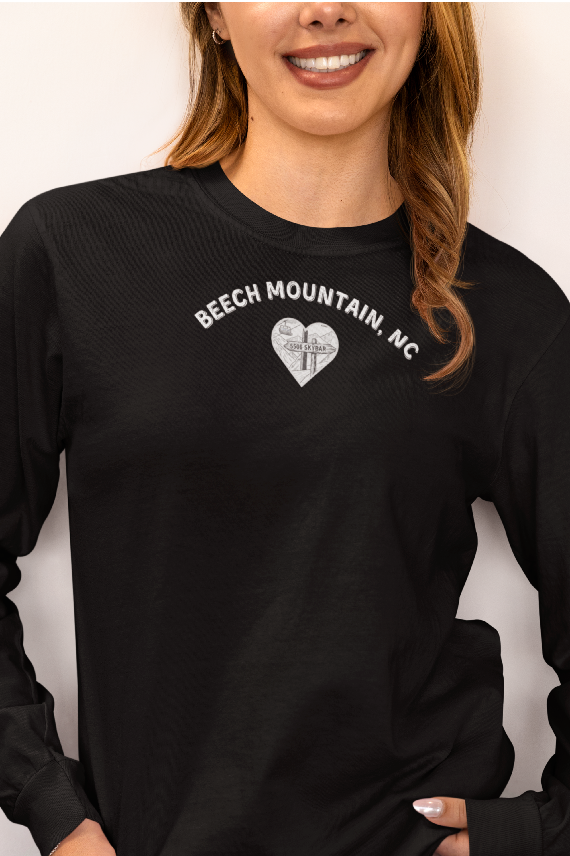 Woman wearing a black Comfort Colors® long-sleeve tee with a heart-shaped illustration of Beech Mountain’s slopes, chairlift, and sign pointing to 5506 Skybar 